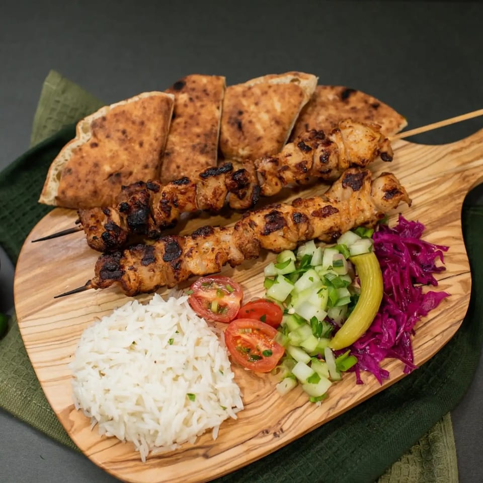 Two chicken shish kebabs, rice, salad, and pita bread on a wooden board.