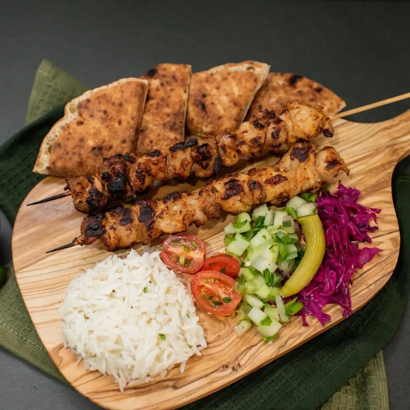 Two chicken shish kebabs, rice, salad, and pita bread on a wooden board.