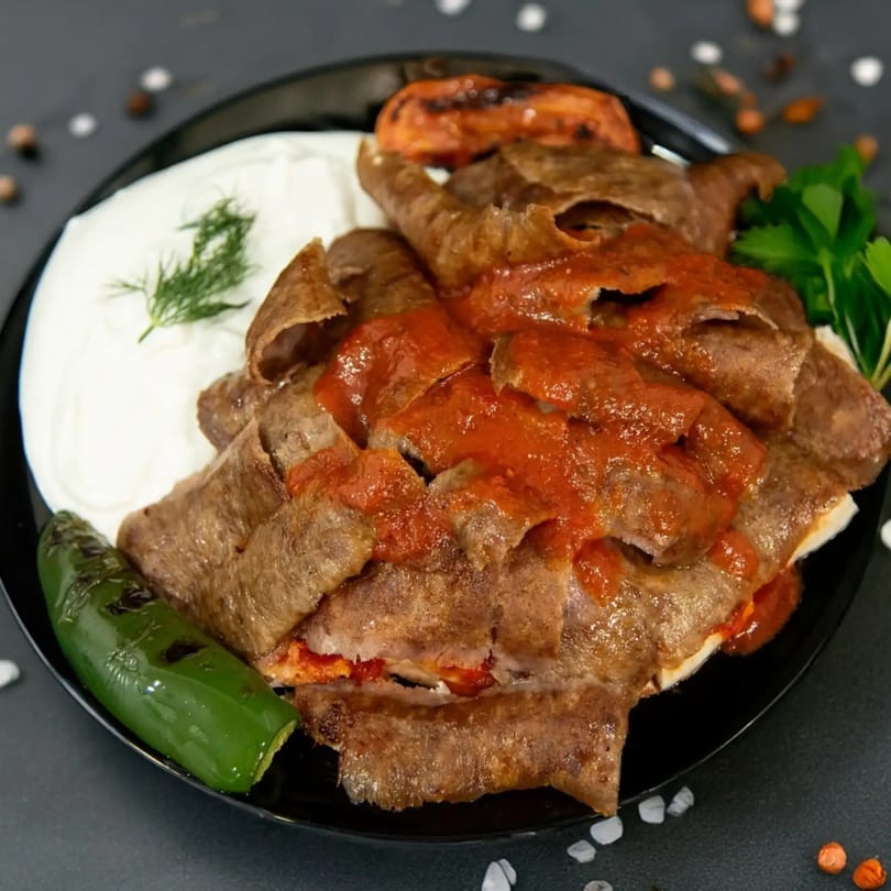 Iskender kebab with tomato sauce, yogurt, grilled tomato, and green pepper on a black plate.
